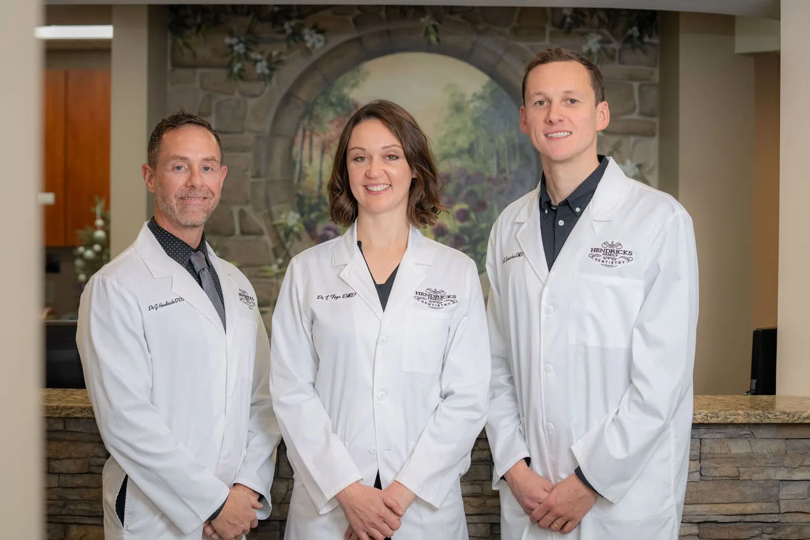 Three dentists in white coats posing and smiling in a dental office with a painted stone arch background.